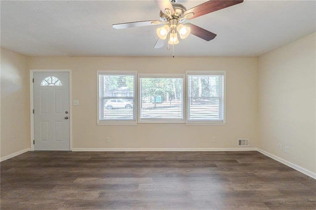 Living room, shown empty, with three windows, a ceiling fan and the front door